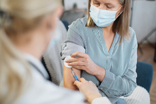 View From The Shoulder Of Female Unrecognizable Doctor. Senior Lady In Protective Mask, Being Vaccinated At Home By Her Family Gp Doctor.