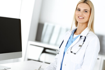 Blond female physician is standing at her workplace near desktop computer. Woman-doctor is excited and happy of her profession. Medicine concept