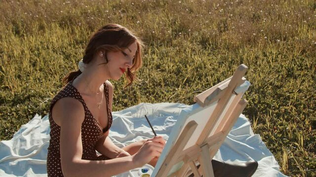 Giovane ragazza dipinge un quadro su tela seduta su una tovaglia bianca in un prato