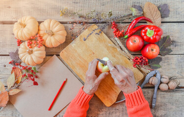Senior woman hands cutting vegetables, cooking process, family tradition. Autumn background, fallen leaves, fruits, wooden table, vintage paper, copy space. Vegan, healthy, Thanksgiving, top view.