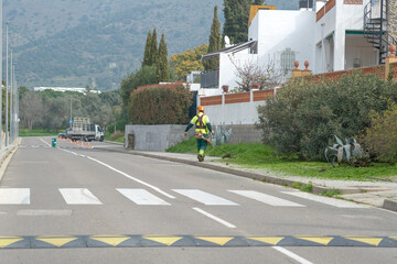 Worker in protective clothing with mowing tall grass with petrol lawn trimmer along the roadway. Public services of the city. Pedestrian crossing, crosswalk. Speed bump, traffic cones.