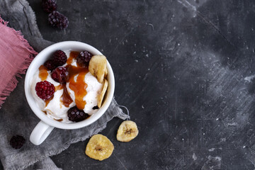 Coconut vegan ice cream in a white cup. Sundae balls and caramel. Ice cream with berries and raspberries. Dark background, copy space.