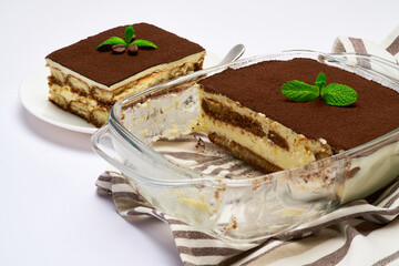 Traditional Italian Tiramisu dessert in glass baking dish, portion on plate and scapula isolated on white background