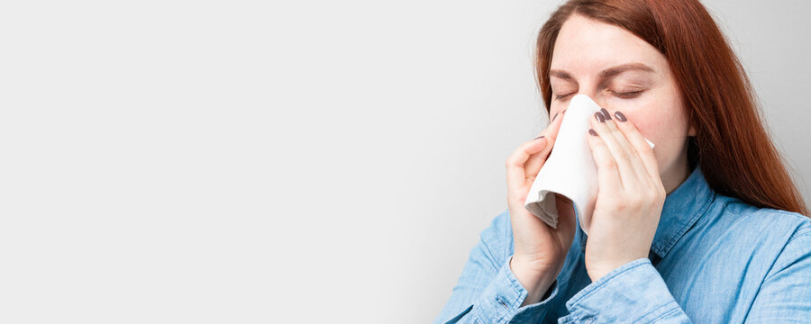 Young Sick Woman Sneezing Holding Napkin Blowout Runny Nose On A Gray Background With Place For Text.