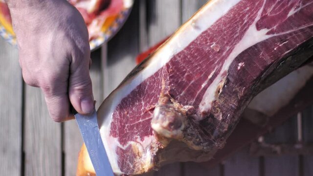 High angle view of man hand cutting a slice of dried spanish jamon serrano on wood table and plate in the background.