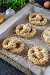 Homemade unbaked soft pretzels with sesame seeds ready to go to the oven in a baking tray. Gluten free dough for savory delicious pastry with a knot. Egg wash, garlic, and coriander in the background