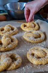 Homemade unbaked soft pretzels ready to go to the oven on a baking sheet. Woman's hand sprinkle sesame on to of pretzels in the background. Gluten free dough for savory delicious pastry with a knot