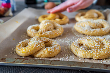 Close up on homemade unbaked soft pretzels ready to go to the oven on a baking sheet. Gluten free dough for a savory delicious pastry with a knot. Selective focus, woman's hand in the background.
