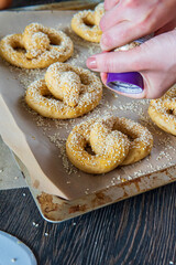 Woman's hand sprinkles garlic flakes on top of homemade unbaked soft pretzels that are ready to go to the oven on a baking sheet. Gluten free dough for savory delicious pastry with a knot. Side view.