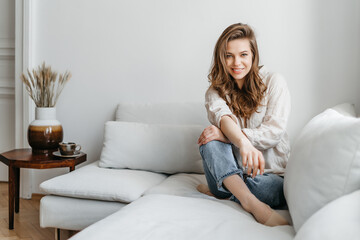 Нoung woman is sitting on the sofa in the living room. A beautiful blonde in white comfortable home clothes.