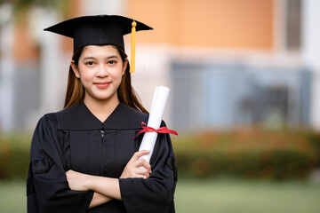 A young happy Asian woman university graduate in graduation gown and mortarboard holds a degree...