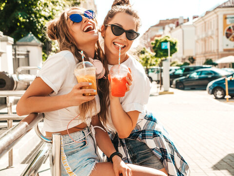 Two Young Beautiful Smiling Hipster Female In Trendy Summer Clothes. Carefree Women Posing Outdoors.Positive Models Holding And Drinking Fresh Cocktail Smoothie Drink In Plastic Cup With Straw