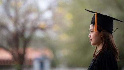 A young happy Asian woman university graduate in graduation gown and mortarboard holds a degree...