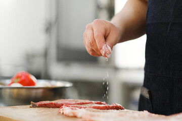 The chef salts and peppers a piece of meat. Cooking in the kitchen of restaurant.