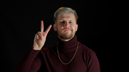 Friendly happy stylish young man with blue eyes in trendy clothes looking at camera, feeling happiness, fun, enjoyment, showing victory sign, hoping for success and win, doing peace gesture, smiling