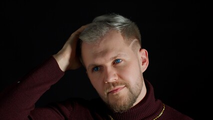 Smiling cheerful young man in trendy stylish clothes holding hand over head, looking at camera on black background. Fashion people sincere emotions. Attractive guy studio shot. Fun and enjoyment