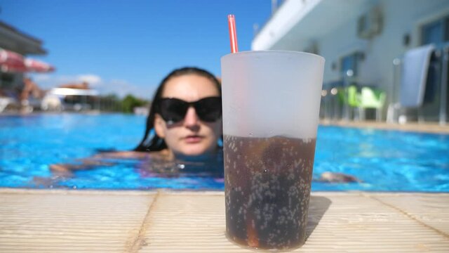 Young sexy girl swimming to edge of pool and quenching thirst with cool drink in hot sunny day. Attractive tanned woman floating in basin of hotel and enjoying resting on resort. Close up Slow motion