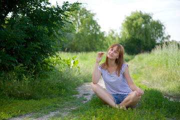 Naklejka na ściany i meble A young girl is sitting on the grass in a meadow. The girl has a rest in nature. Happiness and joy in the fresh air in summer