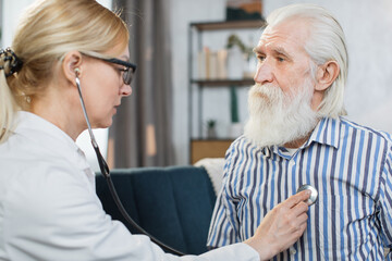 Close up of senior bearded man sitting on the couch, while caring confident blond female doctor listening his heartbeat during home visit.