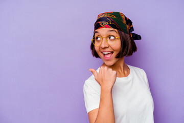 Young mixed race woman wearing a bandana isolated on purple background points with thumb finger away, laughing and carefree.
