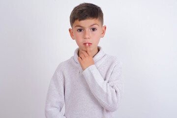 Nervous puzzled Caucasian kid boy wearing knitted sweater against white wall opens mouth from surprise, reacts on sudden news.