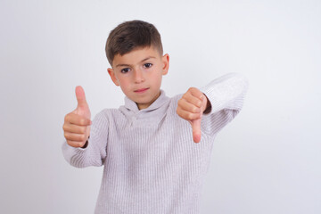 Caucasian kid boy wearing knitted sweater against white wall showing thumbs up and thumbs down,...