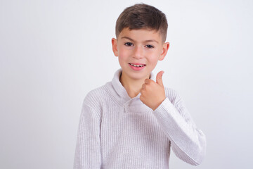 Caucasian kid boy wearing knitted sweater against white wall doing happy thumbs up gesture with hand. Approving expression looking at the camera showing success.