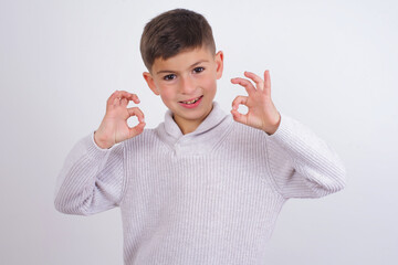 Caucasian kid boy wearing knitted sweater against white wall showing both hands with fingers in OK...