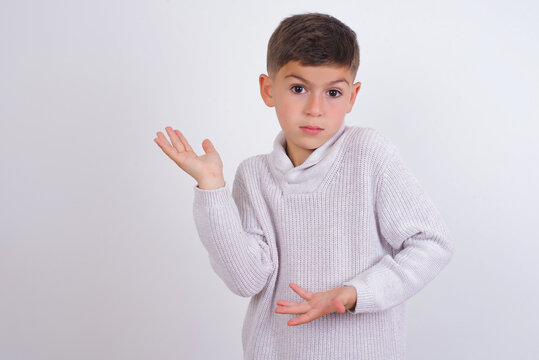 Caucasian kid boy wearing knitted sweater against white wall pointing aside with both hands showing something strange and saying: I don't know what is this. Advertisement concept.