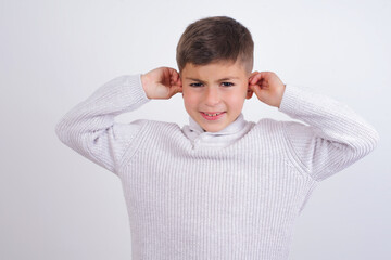 Caucasian kid boy wearing knitted sweater against white wall covering ears with fingers with annoyed expression for the noise of loud music. Deaf concept.