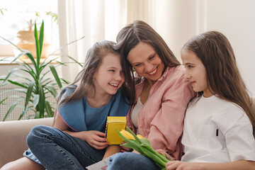 Happy dark-haired young woman embraces her daughters sitting at home on the couch.In their hands is a bouquet of yellow flowers and a homemade gift.The concept of Mother's Day, March 8 and Birthday.