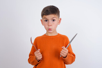Model hungry Caucasian kid boy wearing knitted sweater against white wall holding in hand fork...