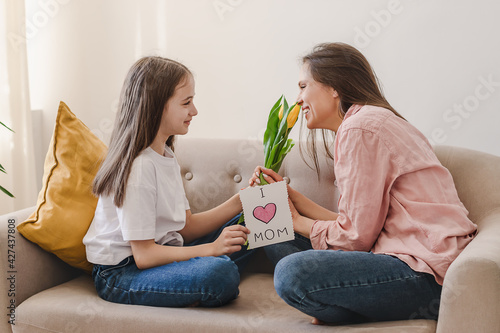 A dark-haired girl congratulates her mother on Mother's Day, gives her bouquet of yellow tulips and homemade greeting card.Home interior design.The concept of Mom's Day,March 8 and Birthday.Copy space