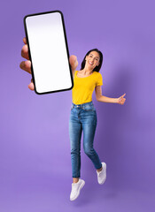 Joyful young woman jumping, gesturing thumbs up and showing mobile phone with empty screen on violet background, mockup