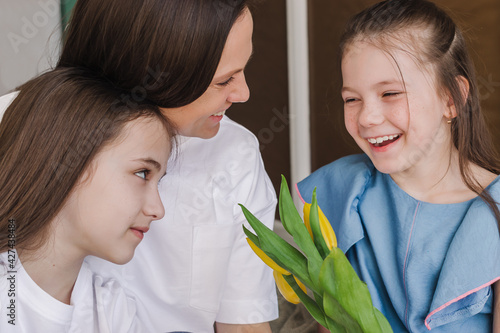 Smiling dark-haired young woman talking with her daughters at home.She is holding a bouquet of yellow flowers.The concept of Mother's Day, March 8 and Birthday.