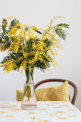 Still life with mimosa blooms in a vase and yellow pillow on a chair