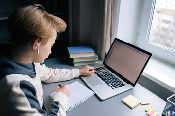 Top back view of pupil boy wearing wireless earphone learning online using laptop making notes in copybook sitting at desk. Child schoolboy doing homework at home. Concept of online distance education