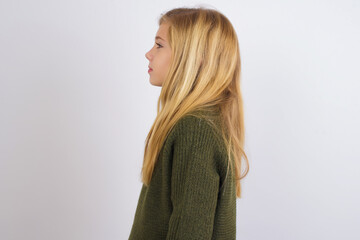 Caucasian kid girl wearing green knitted sweater against white wall looking to side, relax profile pose with natural face with confident smile.