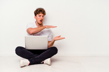 Young caucasian man sitting on the floor holding on laptop isolated on white background shocked and amazed holding a copy space between hands.