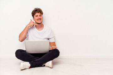 Young caucasian man sitting on the floor holding on laptop isolated on white background showing a mobile phone call gesture with fingers.