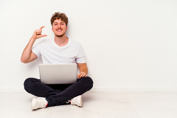 Young caucasian man sitting on the floor holding on laptop isolated on white background holding something little with forefingers, smiling and confident.