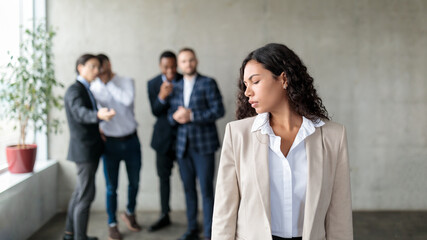 Group Of Businessmen Whispering Behind Back Of Businesswoman In Office