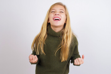 Caucasian kid girl wearing green knitted sweater against white wall celebrating surprised and amazed for success with arms raised and eyes closed. Winner concept.