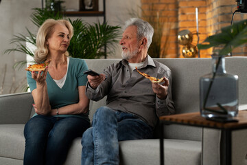 Cheerful husband and wife sitting on sofa at home. Happy senior woman and man eating pizza while watching a movie..