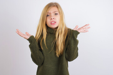 Indignant Caucasian kid girl wearing green knitted sweater against white wall gestures in bewilderment, frowns face with dissatisfaction.