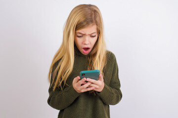 Focused Caucasian kid girl wearing green knitted sweater against white wall use smartphone reading social media news, or important e-mail