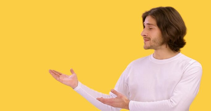 Cheerful millennial man pointing hands aside at empty space and smiling to camera, yellow studio background, slow motion