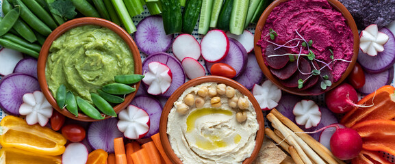 Narrow close up view of three dips surrounded by fresh cut vegetables for dipping. - obrazy, fototapety, plakaty