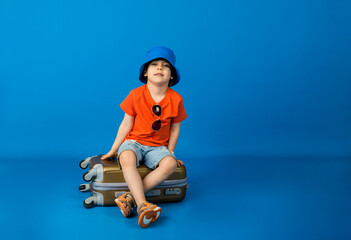 a small boy in an orange T-shirt and denim shorts and a panama hat sits on a suitcase on a blue background with space for text