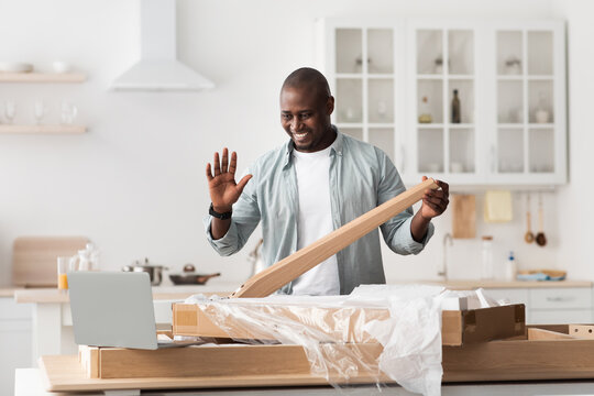 Black handyman shooting video instructions for assembling furniture at home with own hands, waving to laptop at home